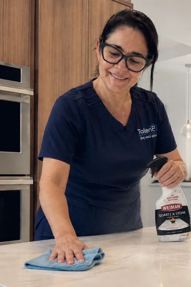 Uma profissional da Talent Cleaning Services passando pano na bancada da cozinha ao lado dos fornos embutidos, com a sala ao fundo.